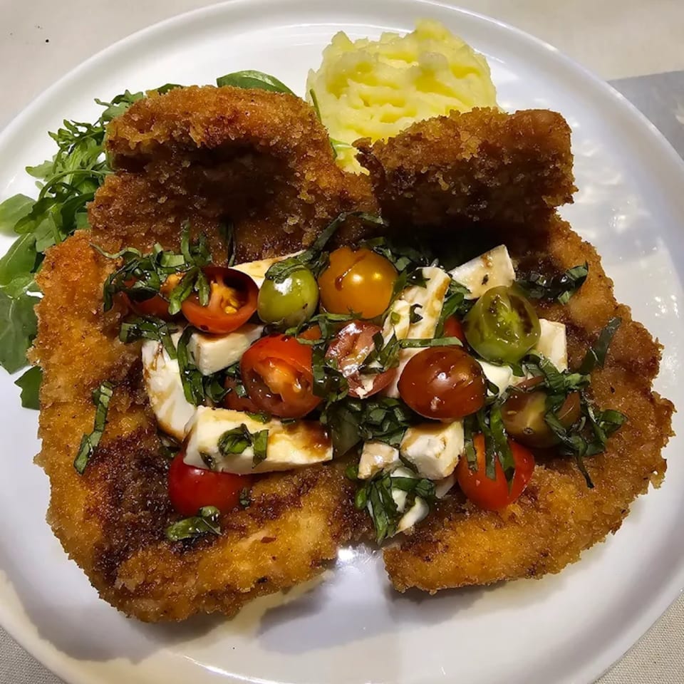 Crispy fried schnitzel topped with colorful cherry tomatoes, mozzarella, and basil, served with mashed potatoes.